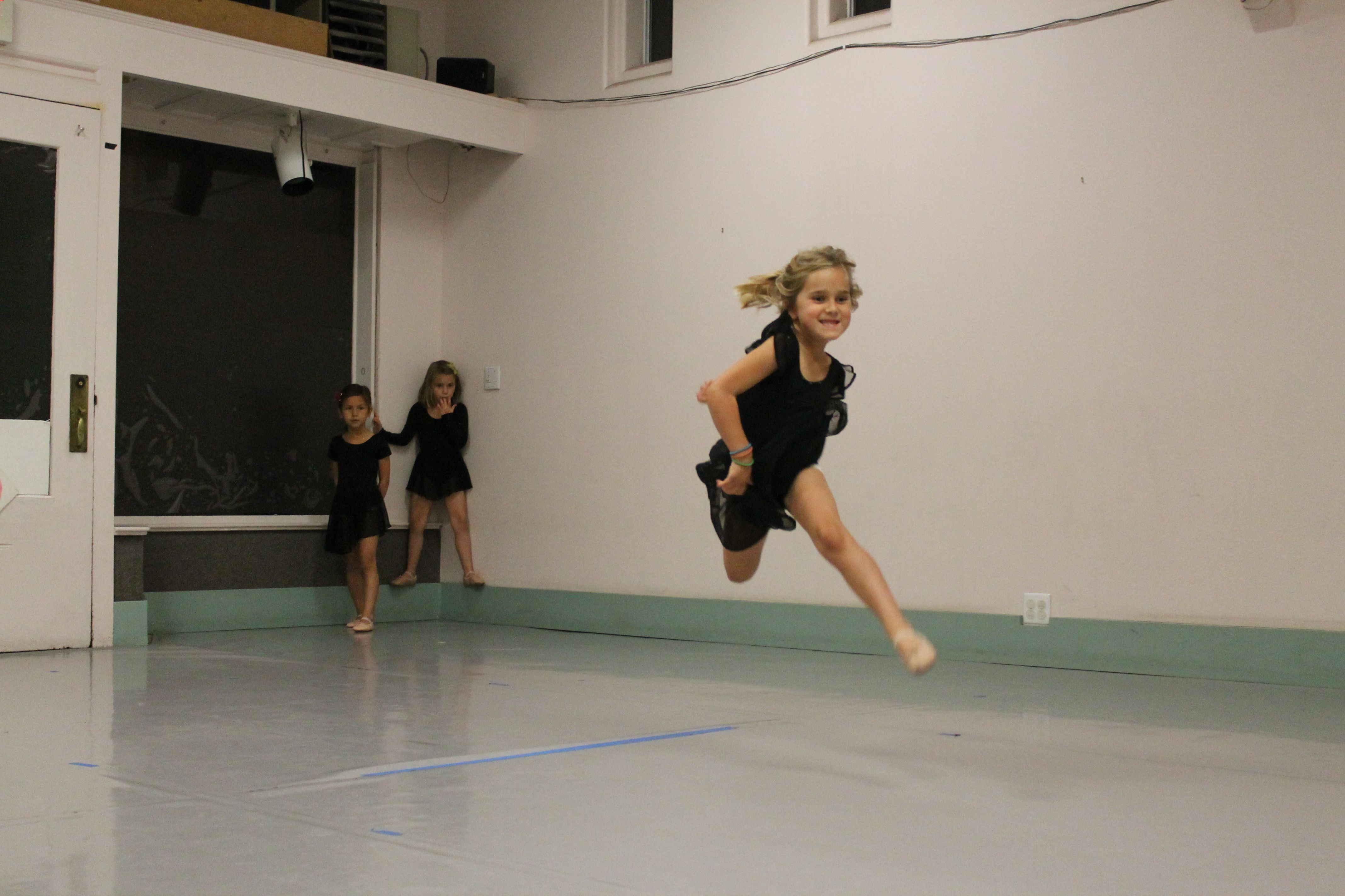 A young dancer leaps during class while two dancers wait their turn