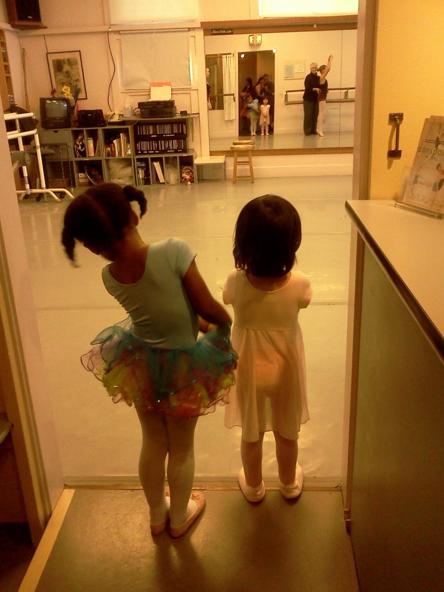 Two young dancers stand in the doorway of the studio watching older students in class