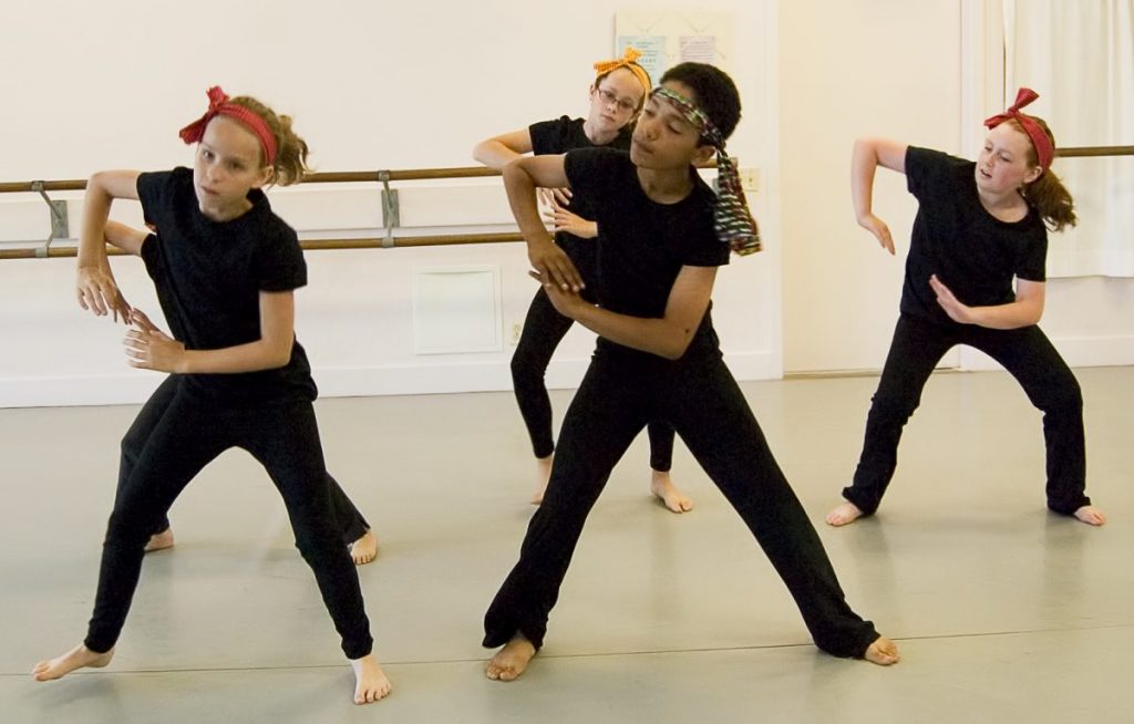4 dancers in all black with colorful head pieces mid rehearsal