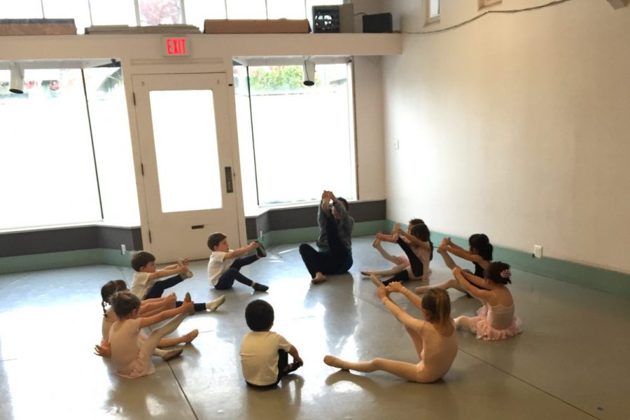 A group of young dancers sit in a circle and stretch with their teacher