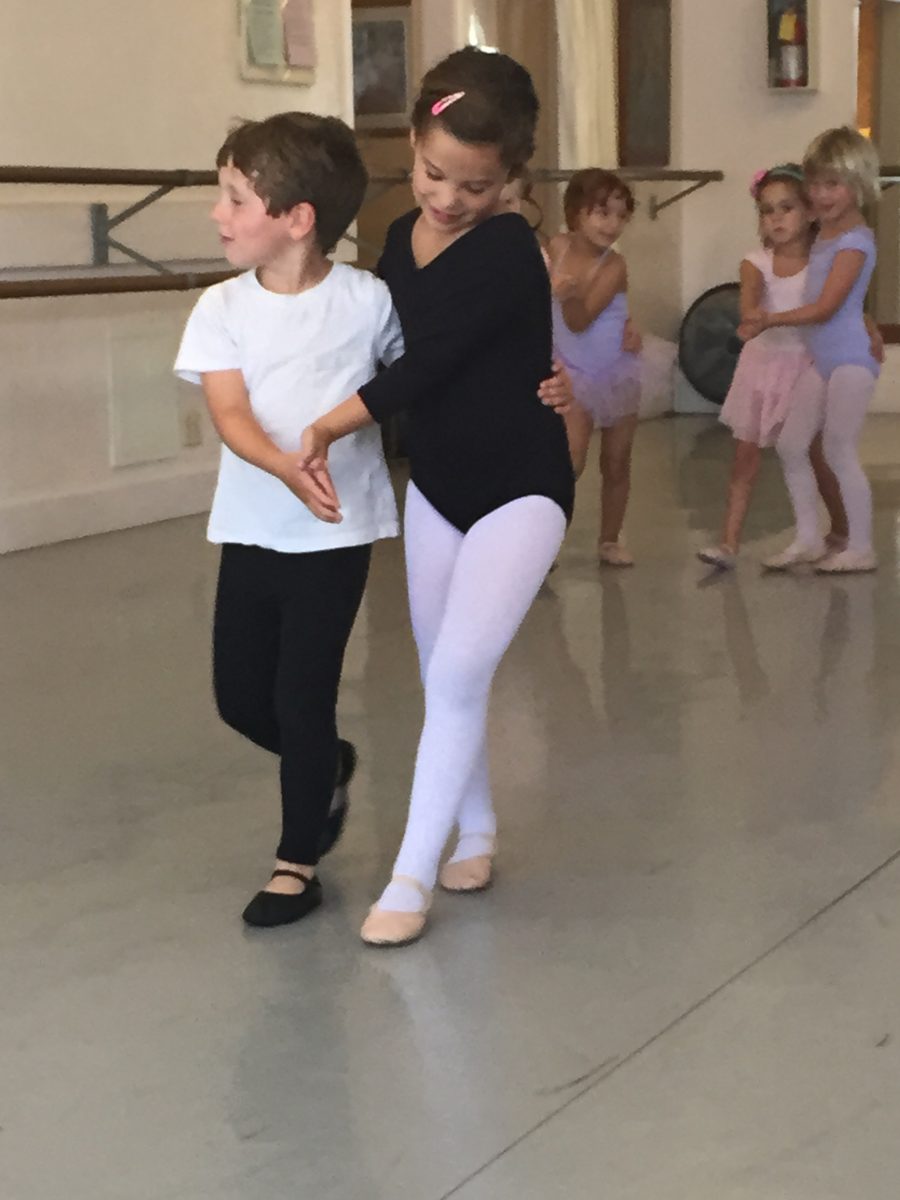 Two young dancers travel across the floor together. Other dancers wait their turn.