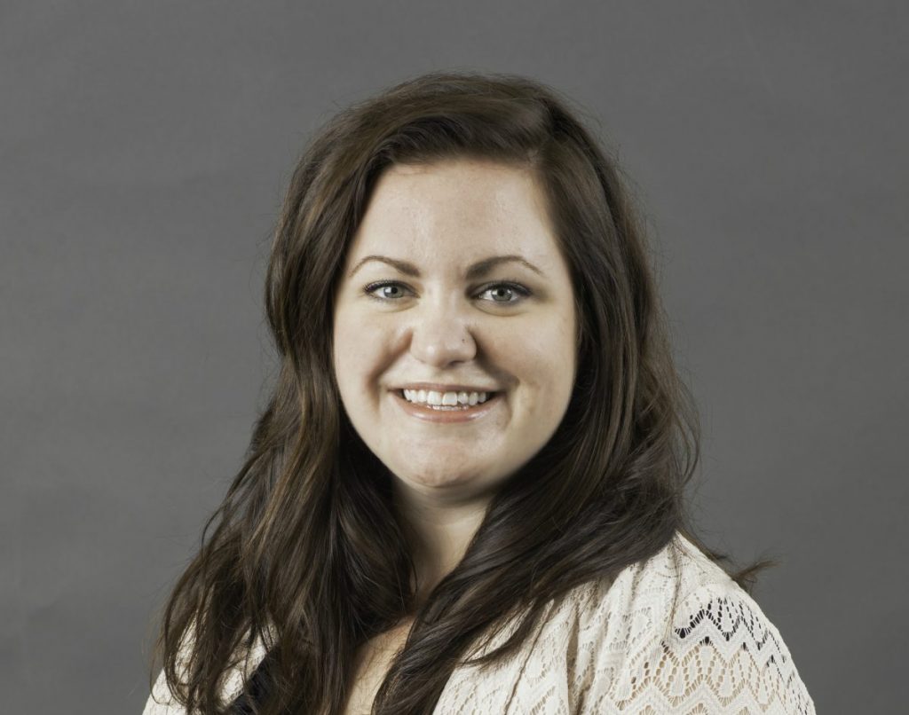 Katie Taylor's headshot. Katie has brown wavy hair and wears a pink lace top.