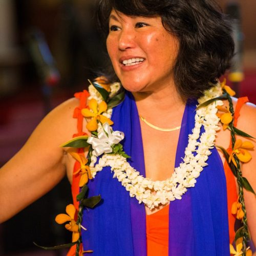 Freesia wears a red top, blue sheer scarf and floral leis. Freesia has dark short hair.
