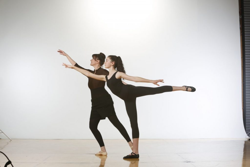 An adult dancer in a low arabesque poses with a teen in a 90 degree arabesque