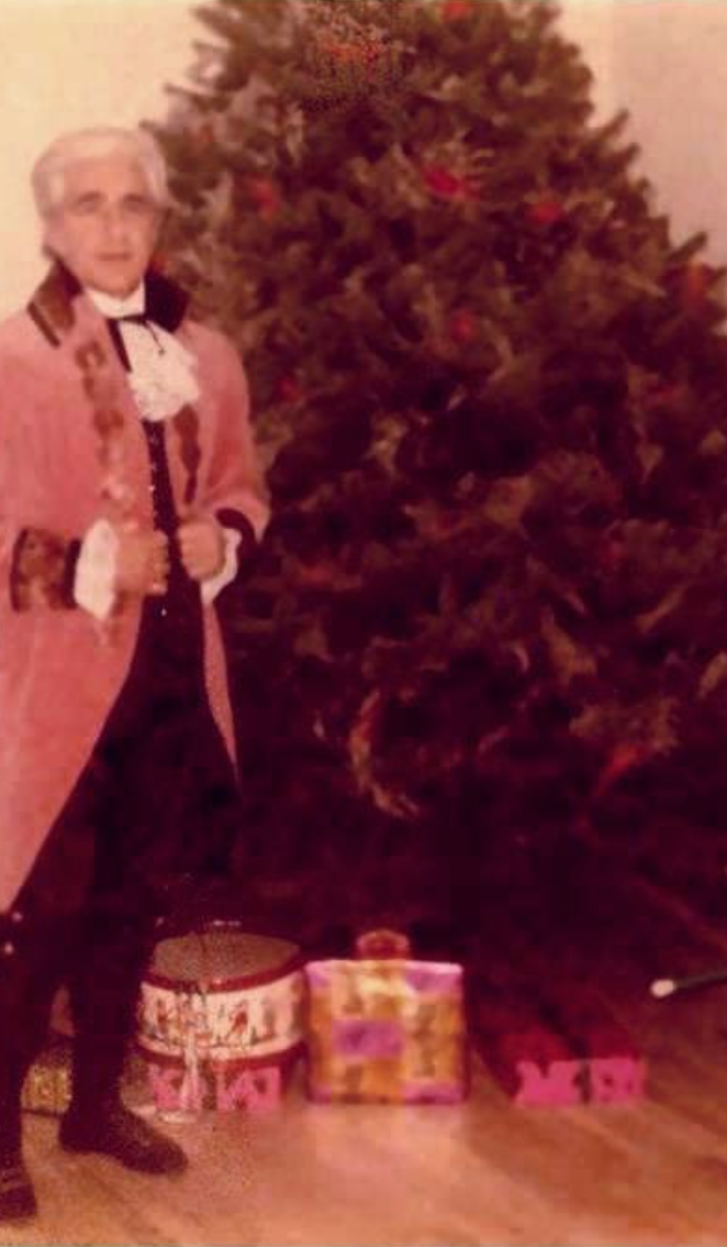 Scan of an old photo of Mr. Corvino in costume standing in front of a Christmas tree.