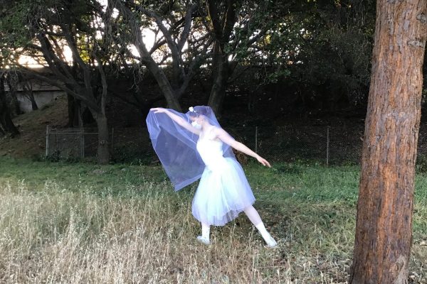 A dancer in costume for Giselle stands in the grass in front of some trees, posed.