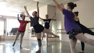 5 dancers mid dance in a low arabesque, arms lifted to the sky, during rehearsal.