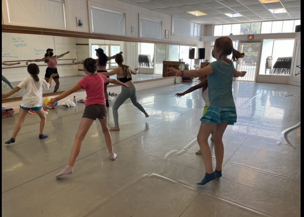 5 students receive instruction from a teacher in the front. Dancers are mid-movement with arms reaching wide.