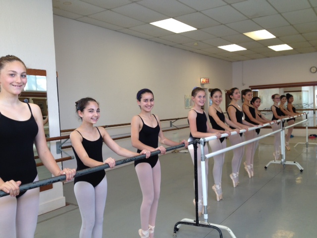11 pre-teen dancers at the barre in their pointe shoes standing en pointe in parallel and smiling at the camera
