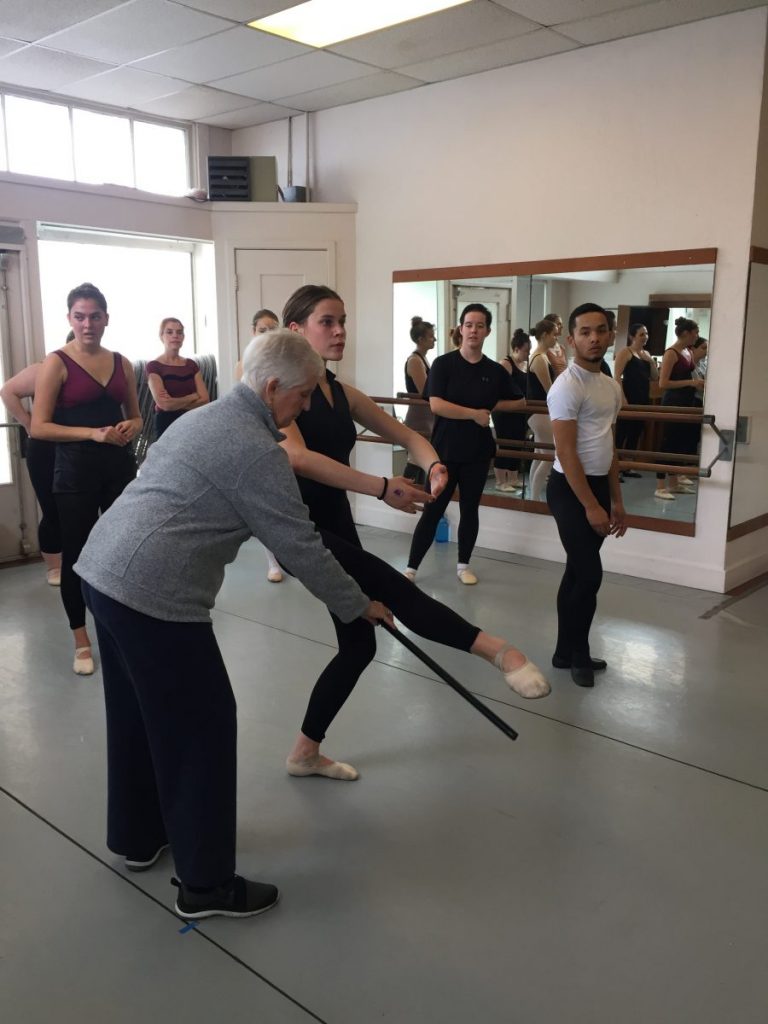 Beth holds a black stick while a dancer demonstrates a leg extended to the front. Other dancers in class observe.
