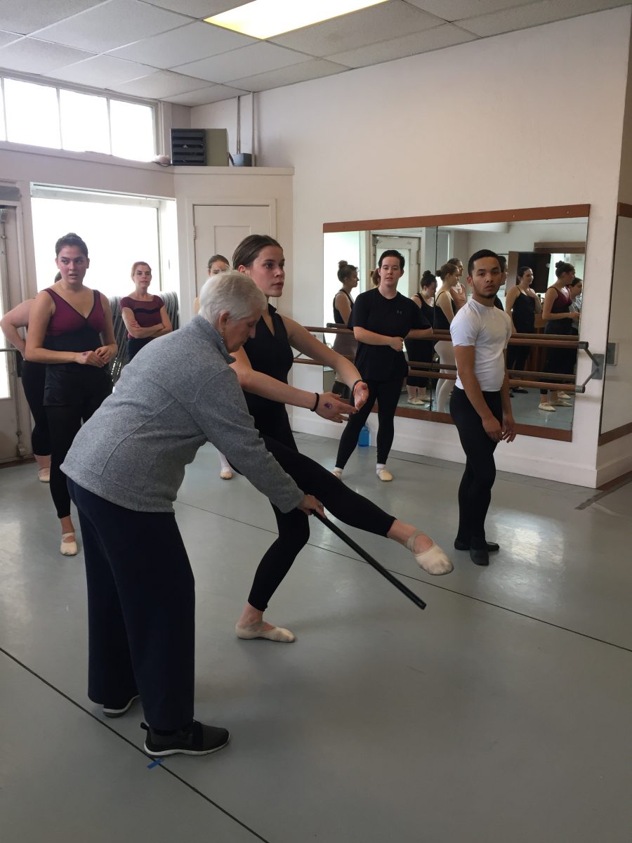 Beth holds a black stick while a dancer demonstrates a leg extended to the front. Other dancers in class observe.